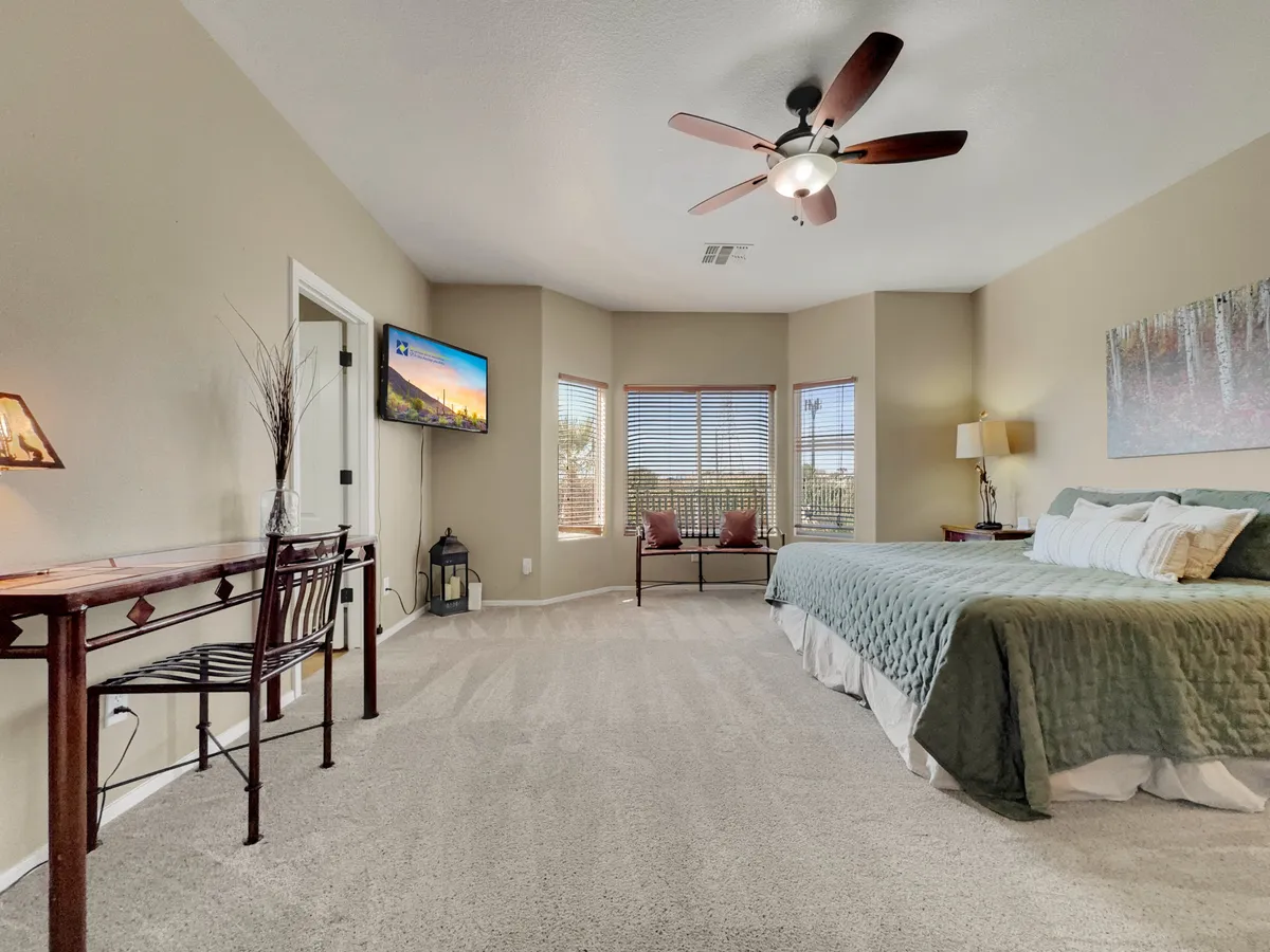 A tranquil bedroom with a large bed dressed in green bedding, a cozy seating area by the window, a television mounted on the wall, and decorative artwork. Soft natural light fills the space, creating an inviting, restful ambiance.