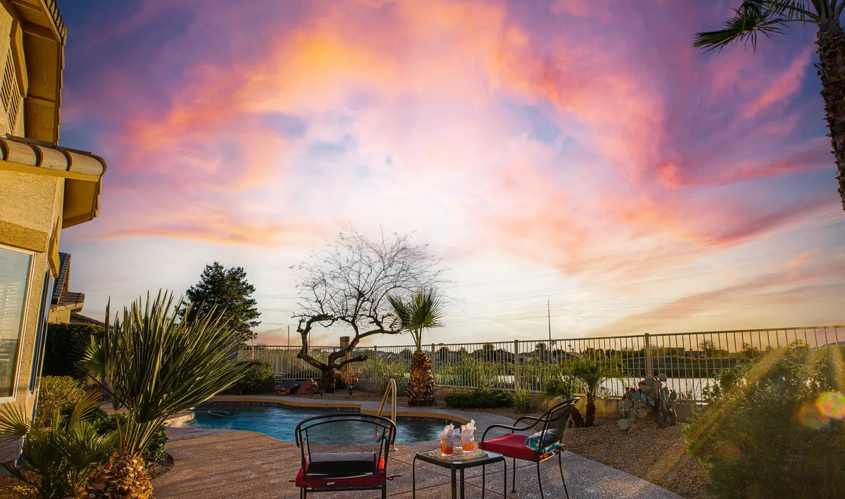 This image shows a serene backyard with a sparkling pool and comfortable seating area, all bathed in the warm glow of a sunset sky. The tranquil ambiance and inviting setup suggest an ideal spot for relaxation and socializing, with the pool reflecting the vibrant sunset colors.