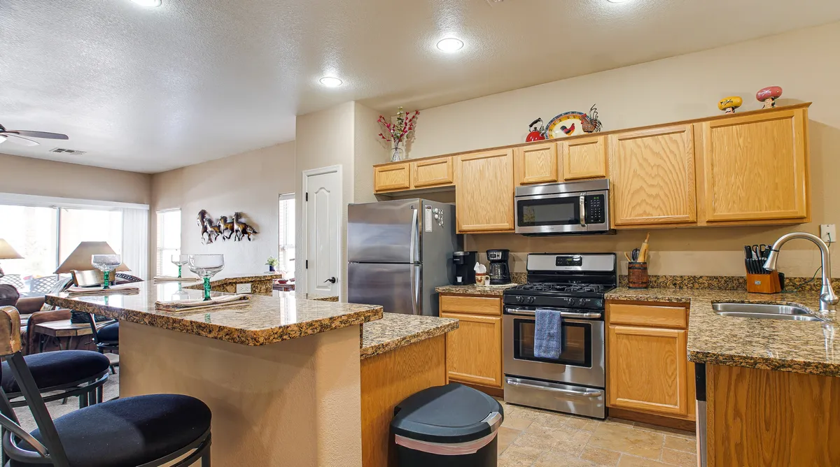 An open kitchen with a granite countertop, stainless steel appliances, wooden cabinets, a central island, and a clear view extending into the living area. The setup promotes easy movement and social connection across spaces.