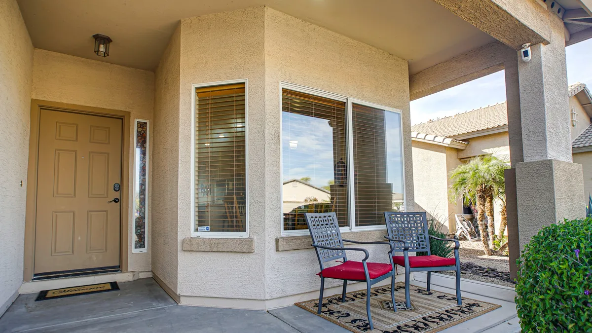 The image captures the inviting front exterior of the home, featuring a welcoming doorway framed by two chairs and a window offering glimpses of the interior. The setting evokes tranquil sunrise coffee moments or evening chats under the stars, suggesting both privacy and a connection to the neighborhood.