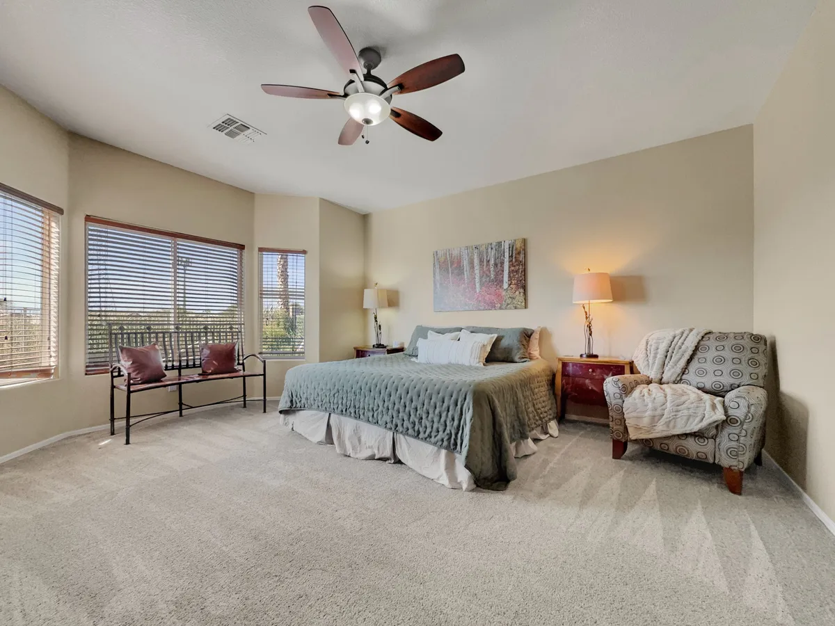 A serene bedroom featuring a bed with a quilted cover, large windows with wooden blinds, a uniquely patterned armchair, a nightstand with a lamp, and framed wall art. The room is bright and airy, with a soothing, cozy atmosphere.