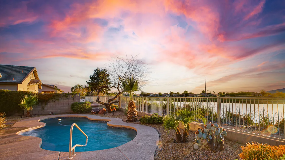 A beautifully landscaped backyard features a shimmering swimming pool set beside a tranquil water body, framed by lush trees and illuminated by a vibrant sunset sky. Comfortable seating and open space invite relaxation and outdoor fun, with the calming lakefront atmosphere in clear view.
